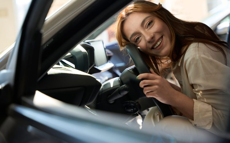 Smiling joyful young Caucasian lady lying in motorcar and hugging steering wheel