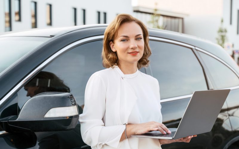 smiling-manager-using-laptop-standing-near-new-car-2022-11-19-02-36-35-utc